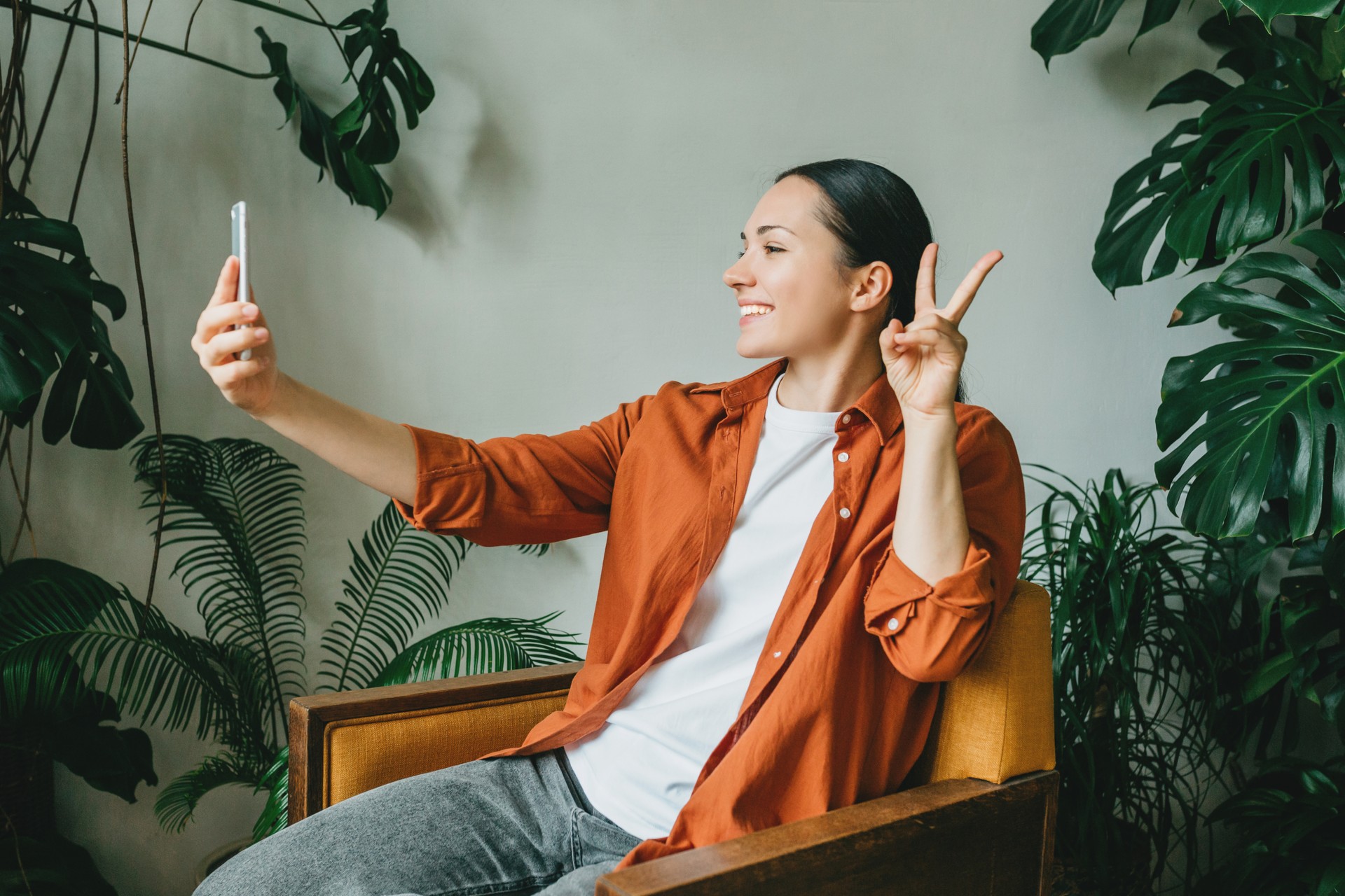 A young smiling woman is sitting at home in a yellow armchair in a green garden and taking selfies, talking on a video connection on the phone.